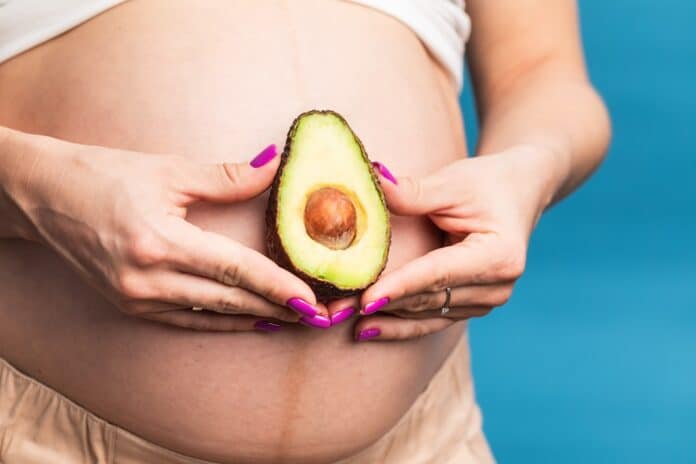 Close-up of a pregnant belly with an avocado held by hands against a blue background AVOCADO