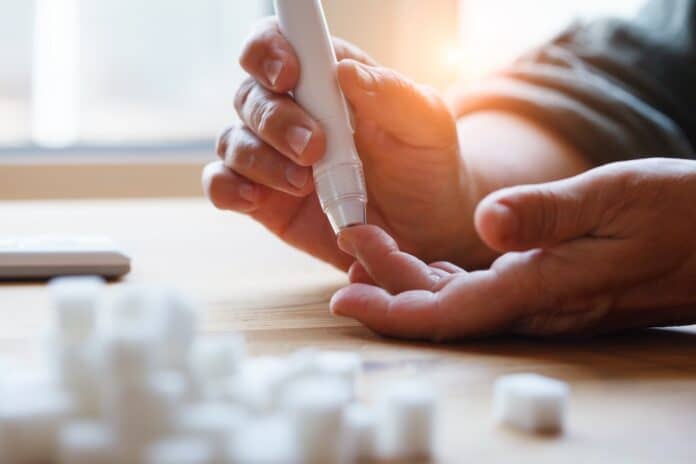 senior woman hands using lancet on finger at home to check blood sugar level, glucometer and sugar cubes on wooden table close up, diabetes concept, elderly health care, sunny morning DIABETE