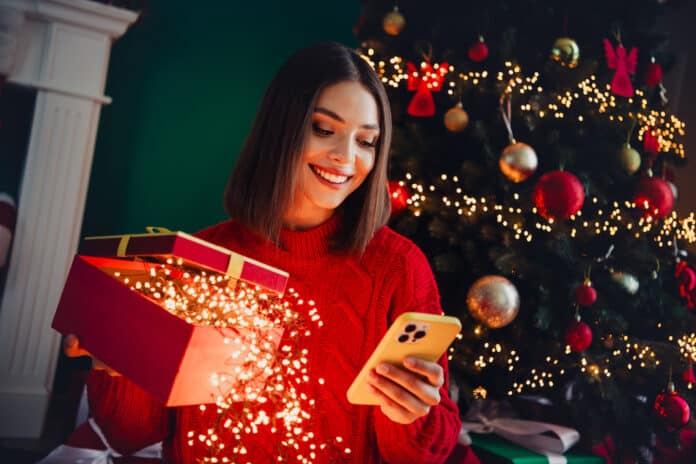 Funny joyful woman in red sweater opens bright gifts beside a twinkling Christmas tree while smiling at her phone in a cozy home scene