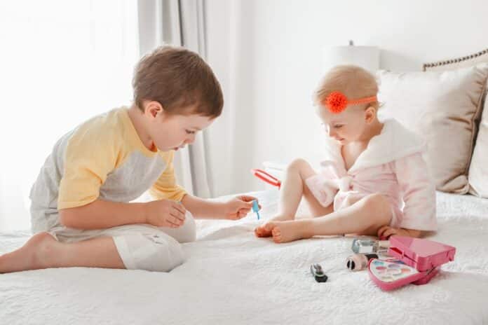 Adorable Caucasian boy and girl siblings playing together painting nails sitting on bed at home. Older brother doing manicure pedicure for his little younger sister. Friends toddlers using cosmetics. SMALTI PER BAMBINI