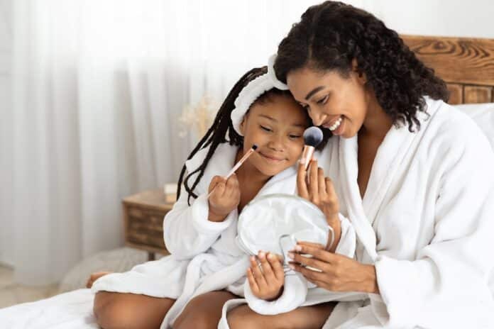 African mother and her little daughter in bathrobes making makeup on bed PFAS