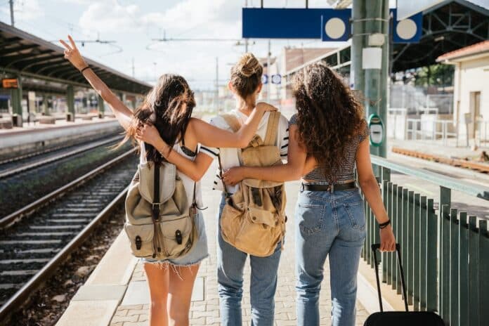 Three young beautiful female women at station to catch train for their vacation together during Coronavirus Covid-19 pandemic wearing protective face masks - Millennials have fun during the holidays interrail