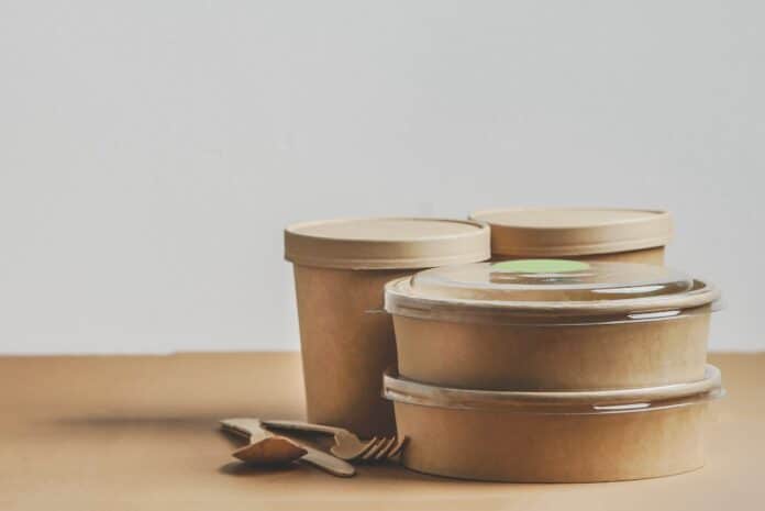 Selection of modern sustainable food delivery containers and cutlery on kitchen table at white wall background. Front view with copy space. Banner PFAS CONTENITORI ALIMENTI