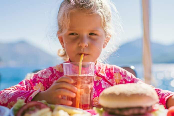 Little girl sipping a soft drink through a straw next to a hamburger at a sunny seaside cafe table FEGATO GRASSO
