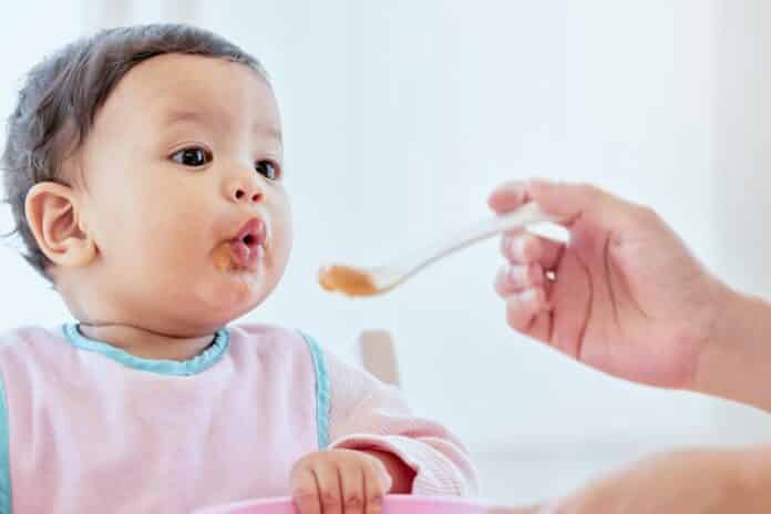 Mother, feeding baby girl and spoon in home, kitchen or living room for nutrition, growth or health. Woman, hand and food for infant child for help, eating or balance in diet for development in house baby food